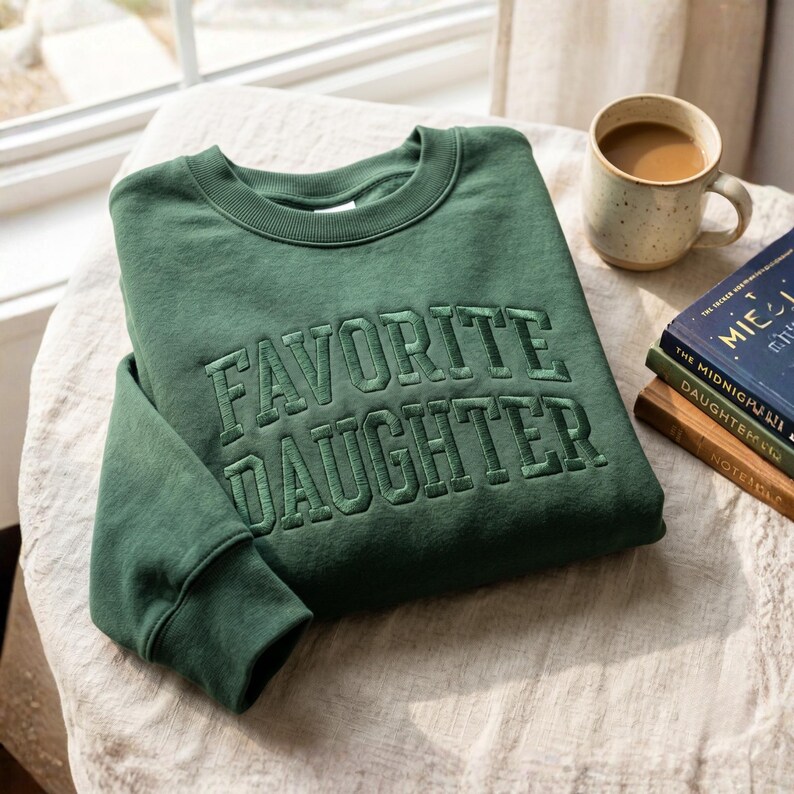 Pode incluir: Um moletom verde floresta com o texto bordado "FAVORITE DAUGHTER". O moletom est&aacute; dobrado sobre uma superf&iacute;cie creme, com uma caneca de caf&eacute; e uma pilha de livros ao fundo.