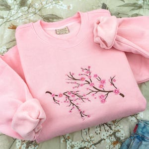 May include: A light pink crewneck sweatshirt featuring an embroidered design of brown branches with small pink cherry blossom flowers. The garment has a ribbed collar and long sleeves with ribbed cuffs. A small brand tag is visible at the neckline.