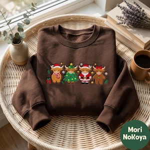 Könnte beinhalten: Ein dunkelbraunes Sweatshirt mit Rundhalsausschnitt und einer Stickerei von vier Highland-Rindern. Die Rinder tragen festliche Kleidung, darunter eine Weihnachtsmannmütze, ein Weihnachtsbaumkostüm, ein Weihnachtsmannkostüm und ein Lebkuchen-Outfit.