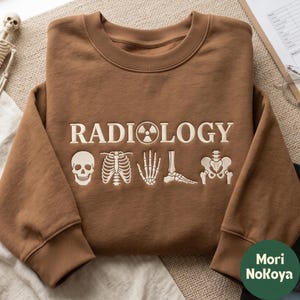 以下が含まれることがあります： 茶色のクルーネックスウェットシャツ。胸元には「RADIOLOGY」の刺繍があり、文字の「O」が放射能マークに置き換わっています。その下には、人間の頭蓋骨、肋骨、手、足、骨盤の白い刺繍イラストが施されています。