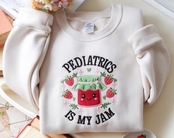 May include: A cream-colored crewneck sweatshirt featuring an embroidered design of a smiling red jam jar with a green lid, surrounded by strawberries and the black text "PEDIATRICS IS MY JAM" arched above and below the central graphic illustration.
