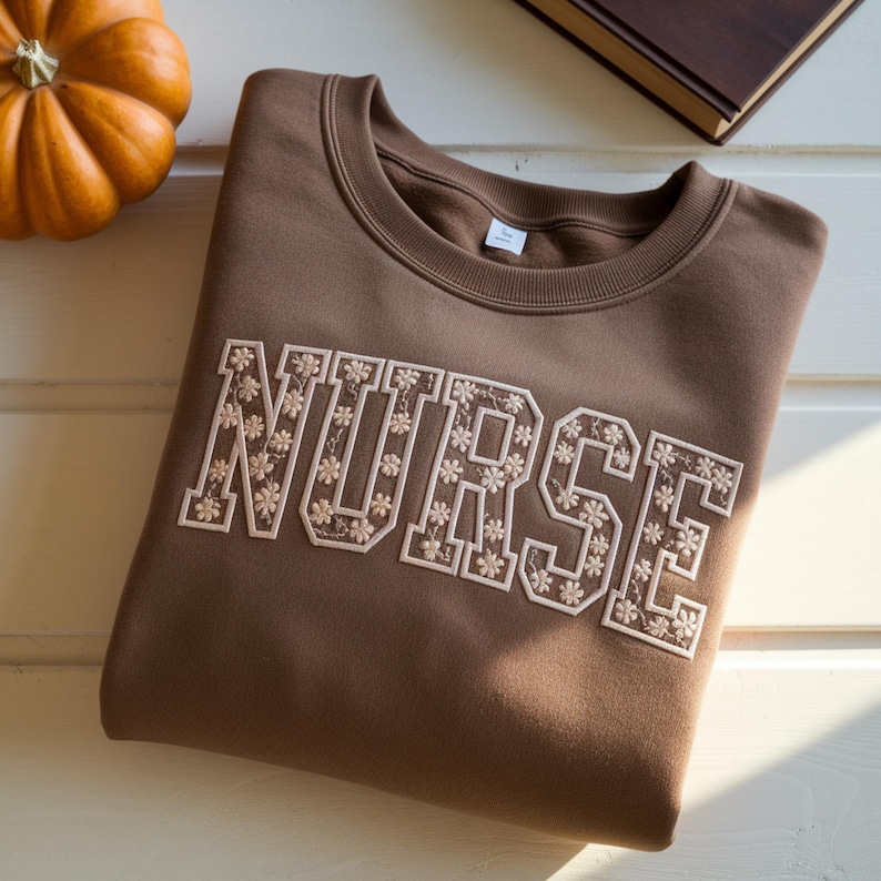 May include: A brown sweatshirt with the word "NURSE" embroidered in a floral design. The sweatshirt is folded on a white surface, with a small pumpkin and a book in the background.