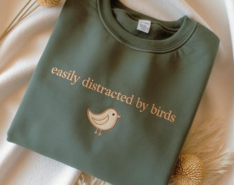 May include: An olive green sweatshirt with the embroidered phrase "easily distracted by birds" and a small bird graphic. The crew neck sweatshirt is folded on a white surface, with decorative dried flowers.