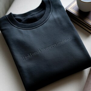 May include: A black crewneck sweatshirt featuring the embroidered text "Hold on, I'm overstimulated." in a matching dark thread color. The garment has a classic ribbed neckline and a smooth, solid-colored fabric finish across the front chest area.