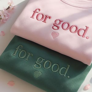 May include: Two sweatshirts, one pink and one green, are stacked. The pink sweatshirt has the embroidered phrase "for good." in pink, with a small heart. The green sweatshirt has the same phrase in green, with a heart.
