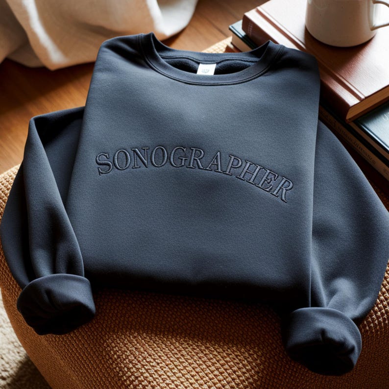 May include: A navy blue sweatshirt with the word "SONOGRAPHER" embroidered across the chest. The sweatshirt has a crew neck and cuffed sleeves. It is resting on a woven surface, with books and a mug in the background.