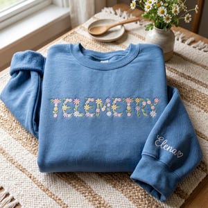 May include: A blue crewneck sweatshirt featuring the word TELEMETRY embroidered in a floral font with pink, yellow, and white flowers and green leaves. The right sleeve cuff features the name Elena embroidered in white cursive script with a small heart.