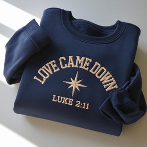May include: Navy blue sweatshirt with the embroidered text "LOVE CAME DOWN" above a starburst design and the text "LUKE 2:11". The sweatshirt has a crew neck and long sleeves.