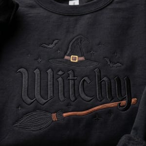 May include: A black sweatshirt featuring an embroidered design with the word Witchy in a gothic font. The design includes a black witch hat with a brown band and gold buckle, a brown broomstick, two small black bats, and several small star icons.