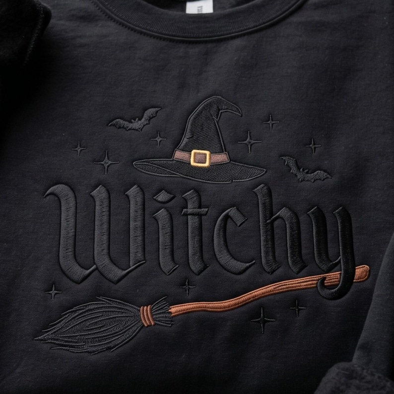 May include: A black sweatshirt featuring an embroidered design with the word Witchy in a gothic font. The design includes a black witch hat with a brown band and gold buckle, a brown broomstick, two small black bats, and several small star icons.