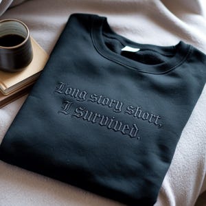 May include: Black crewneck sweatshirt with the embroidered phrase "Long story short, I survived." The sweatshirt is folded and resting on a light-colored surface, with a cup of coffee and a book visible in the background.