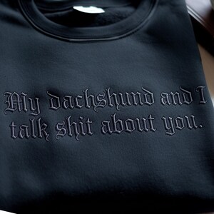 May include: Dark navy sweatshirt featuring the embroidered phrase "My dachshund and I talk shit about you" in a gothic font. The sweatshirt is made of a soft material.