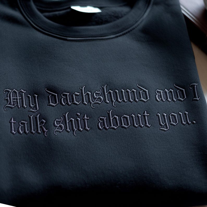 May include: Dark navy sweatshirt featuring the embroidered phrase "My dachshund and I talk shit about you" in a gothic font. The sweatshirt is made of a soft material.