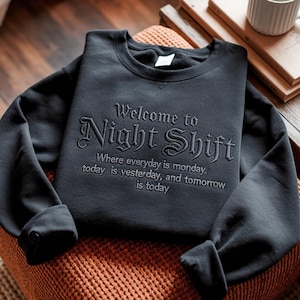 May include: A black crewneck sweatshirt featuring dark embroidery that reads Welcome to Night Shift, Where everyday is monday, today is yesterday, and tomorrow is today. The garment has long sleeves, a ribbed collar, and a soft interior lining.