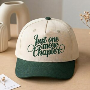 May include: A cream-colored baseball cap with a dark green brim and embroidered dark green text on the front that reads Just one more Chapter in a cursive script font. The cap features a structured crown with a top button and ventilation eyelets.