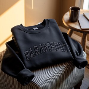 May include: A black crewneck sweatshirt featuring the word PARAMEDIC embroidered in a matching black, arched, collegiate-style font across the chest. The garment has long sleeves with ribbed cuffs and a ribbed neckline, showcasing a clean design.