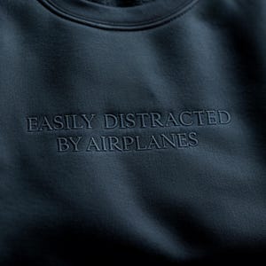 May include: Navy blue sweatshirt with the embroidered text "EASILY DISTRACTED BY AIRPLANES". The crew neck sweatshirt is made of a soft material.