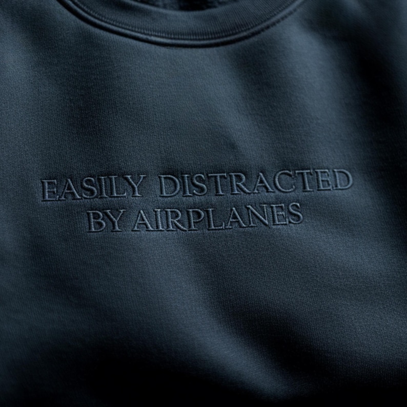 May include: Navy blue sweatshirt with the embroidered text "EASILY DISTRACTED BY AIRPLANES". The crew neck sweatshirt is made of a soft material.