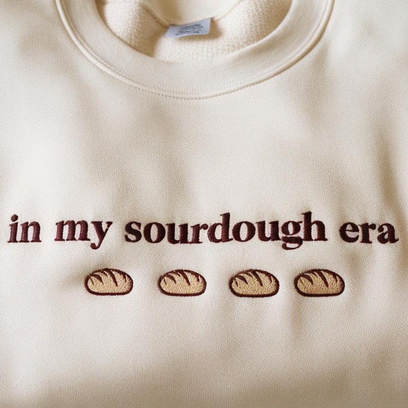 Może przedstawiać: Kremowa bluza z haftowanym napisem "in my sourdough era" w kolorze ciemnobrązowym. Poniżej tekstu znajdują się cztery haftowane wzory bochenk&oacute;w chleba w kolorze brązowym.