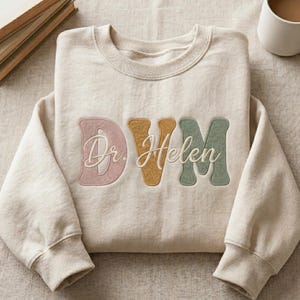 May include: A beige sweatshirt with the embroidered text "Dr. Helen DVM" in pastel colors. The letters "DVM" are in pink, gold, and green, with "Dr. Helen" in a cursive font.