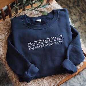 May include: A navy blue crewneck sweatshirt featuring white embroidered text that reads PSYCHOLOGY MAJOR with the phrase Keep talking I'm diagnosing you underneath. The garment has long sleeves and a ribbed collar, cuffs, and hemline.
