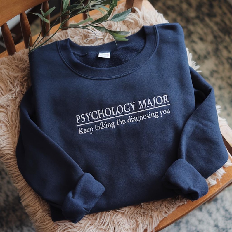 May include: A navy blue crewneck sweatshirt featuring white embroidered text that reads PSYCHOLOGY MAJOR with the phrase Keep talking I'm diagnosing you underneath. The garment has long sleeves and a ribbed collar, cuffs, and hemline.