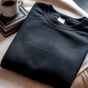 May include: Black crew neck sweatshirt with the embroidered text "cool grandmas club". The sweatshirt is folded on a beige surface, with a cup of coffee and a book in the background.