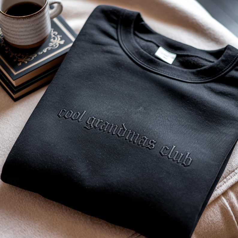 May include: Black crew neck sweatshirt with the embroidered text "cool grandmas club". The sweatshirt is folded on a beige surface, with a cup of coffee and a book in the background.