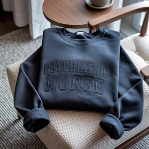 May include: A navy blue sweatshirt with the words "PSYCHIATRIC NURSE" embroidered in a collegiate style. The sweatshirt is folded on a chair with a neutral-toned patterned seat.