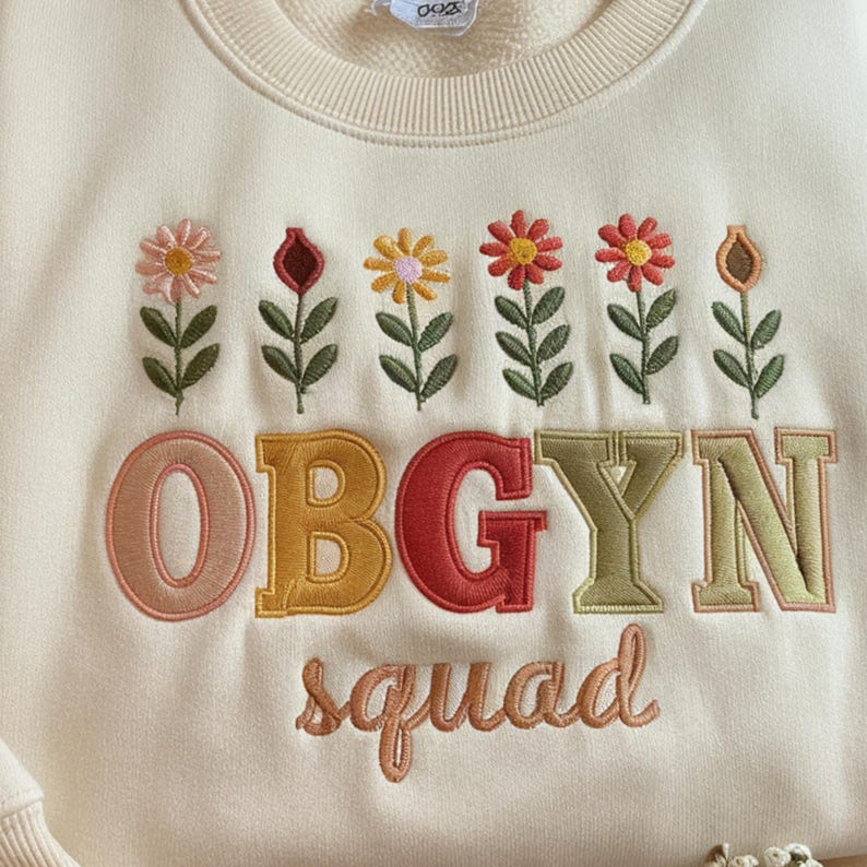 May include: Cream-colored sweatshirt with embroidered floral designs and the text "OBGYN squad." The flowers are in shades of pink, red, yellow, and orange, with green leaves. The letters are in various colors, including peach, yellow, and red.