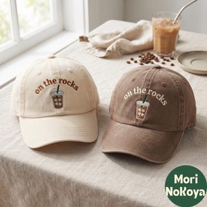 Puede incluir: Dos gorras de béisbol, una en color crema y otra en marrón, cada una con un gráfico bordado de un café helado y el texto "on the rocks" en una fuente curva. Las gorras tienen una corona estructurada, una visera curva y ojales cosidos.