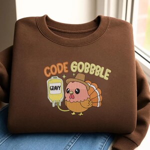 May include: A brown crewneck sweatshirt featuring an embroidered graphic of a turkey wearing a hat, holding a yellow IV bag labeled GRAVY, with the text CODE GOBBBLE in orange and light yellow letters above the turkey and a small star icon.