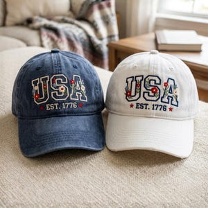 May include: Two baseball caps, one navy blue and one white, are displayed. Both caps feature the embroidered word "USA" with floral accents and the text "EST. 1776". The caps are sitting on a neutral-colored surface.