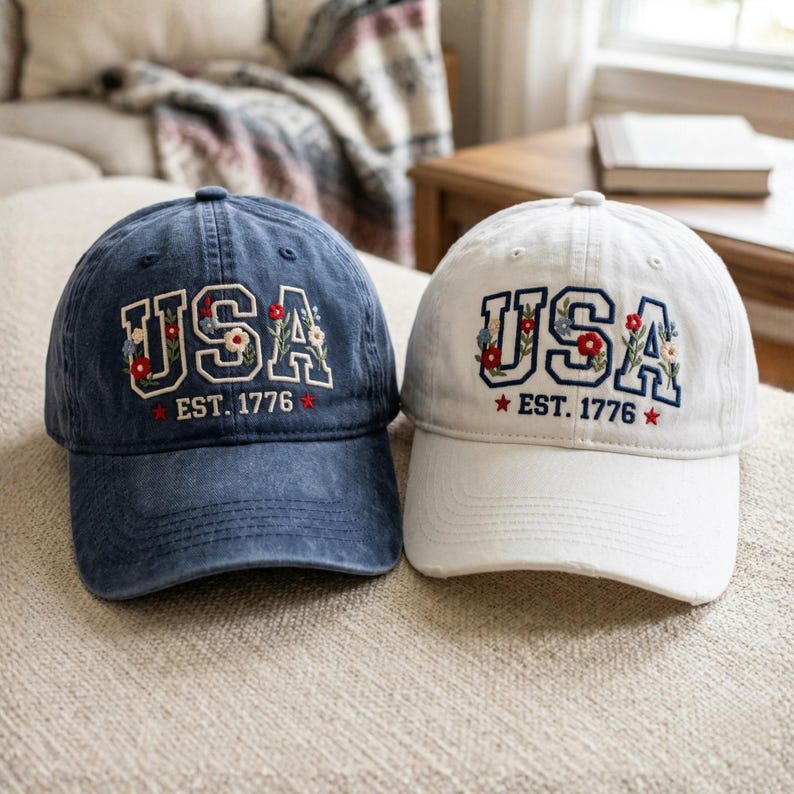 May include: Two baseball caps, one navy blue and one white, are displayed. Both caps feature the embroidered word "USA" with floral accents and the text "EST. 1776". The caps are sitting on a neutral-colored surface.
