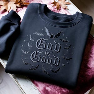 K&ouml;nnte beinhalten: Ein schwarzes Sweatshirt mit Rundhalsausschnitt und einer dunklen Ton-in-Ton-Stickerei auf der Brust. Das Design zeigt den Text "God is Good" in einer gotischen Schrift, umgeben von einer kreisf&ouml;rmigen Anordnung kleiner gestickter Flederm&auml;use und Sterne.
