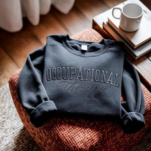 May include: A dark charcoal gray crewneck sweatshirt featuring the text OCCUPATIONAL Therapy embroidered in a matching thread color across the chest. The garment has long sleeves with ribbed cuffs and a classic round neckline design.