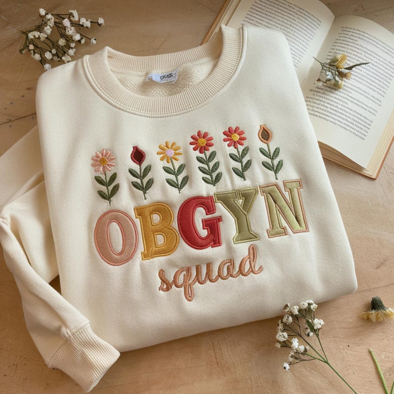 May include: Cream-colored sweatshirt with embroidered floral design and the text "OBGYN squad" in colorful letters. The crew neck sweatshirt has long sleeves and ribbed cuffs. The flowers are in shades of pink, red, yellow, and orange.