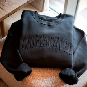 May include: A black long-sleeve crewneck sweatshirt featuring the word EDUMACATED embroidered in a matching black thread across the chest in a bold, arched collegiate-style font. The garment has a ribbed neckline and cuffs.