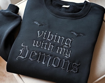 Può includere: Una felpa nera a maniche lunghe con testo ricamato in grigio scuro che recita Vibing with my Demons in un carattere gotico. Due piccole sagome di pipistrello ricamate sono posizionate sopra il testo sul petto del capo.
