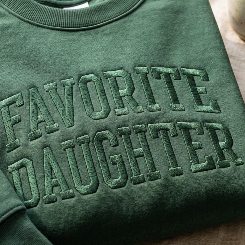Pode incluir: Um moletom verde floresta com o texto bordado "FAVORITE DAUGHTER" em fio verde correspondente. O moletom de gola redonda &eacute; feito de um material macio.