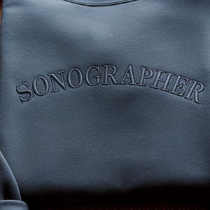 May include: A close-up of a blue-grey sweatshirt with the word "SONOGRAPHER" embroidered across the chest in a matching thread. The text is arched, and the fabric has a subtle texture.
