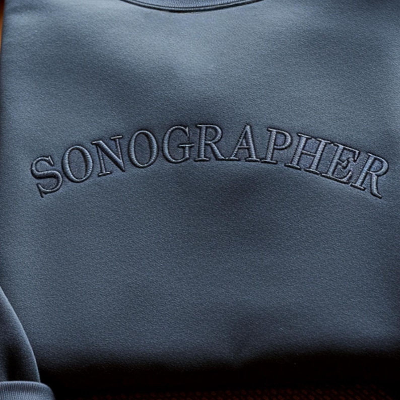 May include: A close-up of a blue-grey sweatshirt with the word "SONOGRAPHER" embroidered across the chest in a matching thread. The text is arched, and the fabric has a subtle texture.