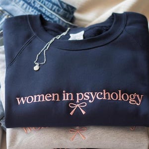May include: A navy blue crewneck sweatshirt featuring the embroidered text "women in psychology" in light pink thread, accompanied by a small pink embroidered bow graphic centered below the text. A silver chain necklace rests on the neckline.