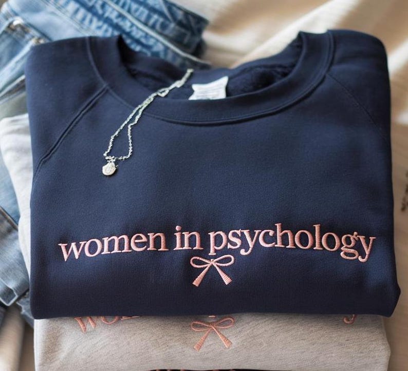 May include: A navy blue crewneck sweatshirt featuring the embroidered text "women in psychology" in light pink thread, accompanied by a small pink embroidered bow graphic centered below the text. A silver chain necklace rests on the neckline.