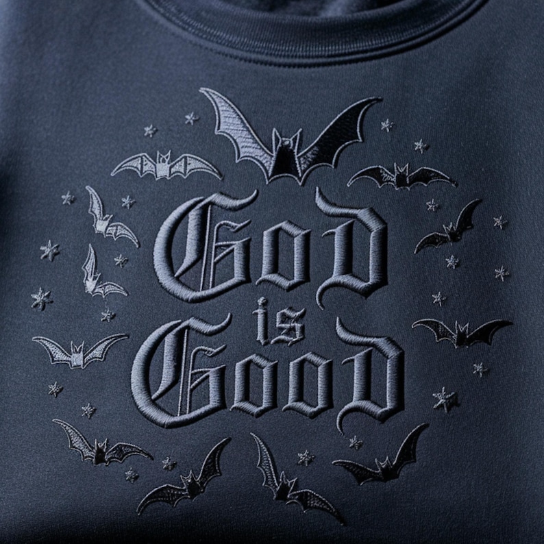 K&ouml;nnte beinhalten: Ein dunkelblaues Sweatshirt mit schwarzer Stickerei. Der Text God is Good ist in einer gotischen Schriftart gehalten und von einem kreisf&ouml;rmigen Muster aus kleinen gestickten Flederm&auml;usen und Sternen umgeben. Das Design ist mittig platziert.