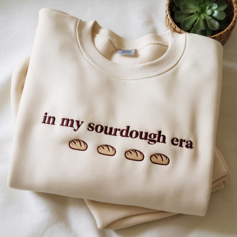 Może przedstawiać: Kremowa bluza z haftowanym napisem "in my sourdough era" brązową nicią. Pod tekstem znajdują się cztery małe, haftowane wzory bochenk&oacute;w chleba. Bluza jest starannie złożona.