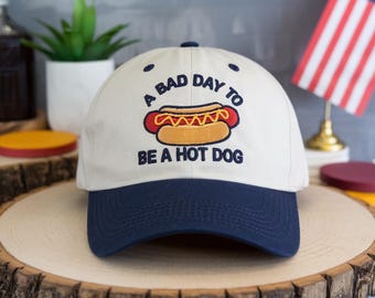 May include: A baseball cap with a white crown and a navy blue brim. The cap features an embroidered hot dog graphic with the text "A BAD DAY TO BE A HOT DOG".