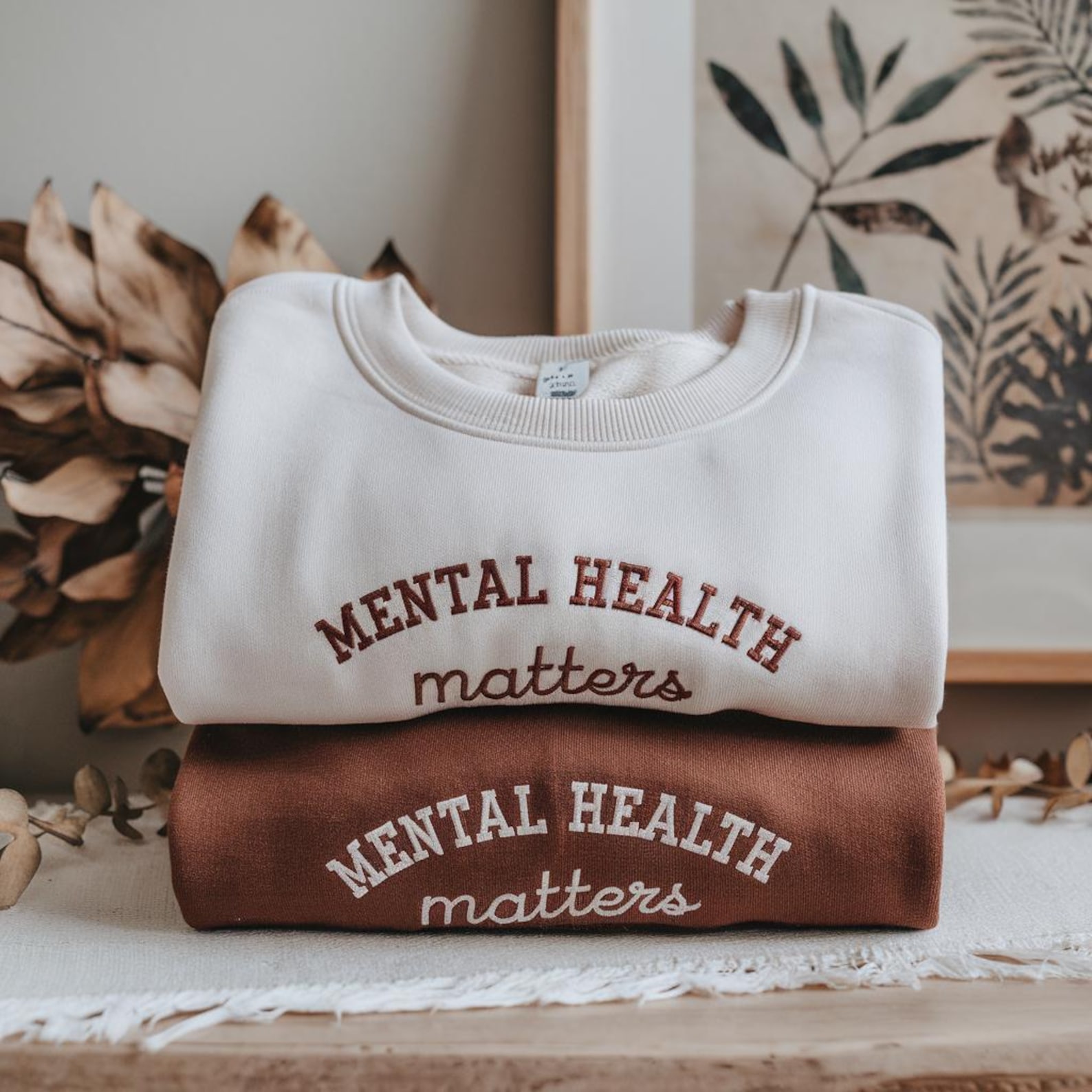 A folded white sweatshirt with brown text on top of a folded brown sweatshirt with white text