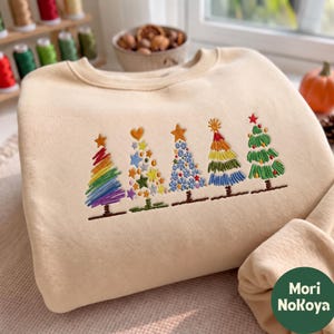 Könnte beinhalten: Ein beigefarbenes Sweatshirt mit Rundhalsausschnitt und einer Stickerei von fünf verschiedenen Weihnachtsbäumen. Die Bäume sind mit bunten Fadenmustern verziert, darunter Regenbogenstreifen, Sterne, Herzen und florale Motive.
