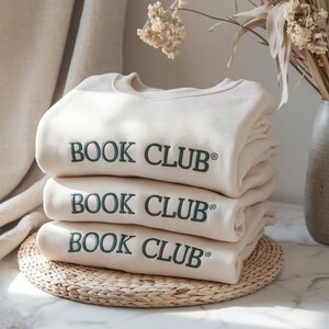 May include: Three cream-colored sweatshirts are stacked on a woven mat. Each sweatshirt has the words "BOOK CLUB" embroidered in dark green across the front. The sweatshirts are folded neatly, suggesting a cozy and organized aesthetic.